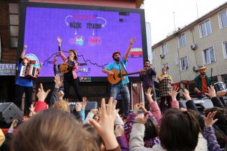 Büyük  bir ekranın önünde konser veren müzisyenler  ve onları izleyen çocukların  elleri yukarıda eşlik ederken görüntüleniyor.