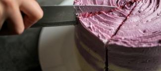 Beyaz bir tabak ve lila rengi aralarda beyaz kreması bulunan yuvarlak bir pastanın yakın plandaki görüntüsü. Bıçak tutan bir el pastada üçgen bir dilim kesmek üzere.