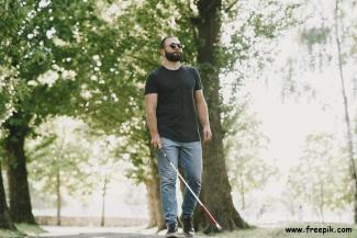 Gündüz vakti yüksek ağaçların olduğu bir parkta tek başına yürüyen beyaz bastonlu siyah gözlüklü, sakallı bir adam. Üzerinde siyah tişört ve mavi bir kot pantolon var. 