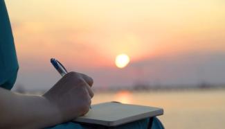 Ufukta kızıl gök   yüzünün arasından görünen güneşe karşı oturarak dizine defteri  dayamış bir kişi sağ eliyle deftere yazı yazıyor. 