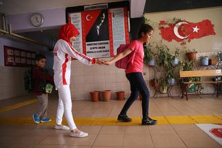 Sarı kılavuz yol döşenmiş bir okul koridoru. Arka planda Atatürk panosu, Türkiye haritası, bir bank, saksı ve çiçekler yer alıyor. Kılavuz yolun üzerinde kırmızı başörtü, kırmızı beyaz tişört ve beyaz pantolon giymiş bir kadın gülümseyerek önünde yürüyen pembe tişört ve pembe sırt çantalı kör kızın elinden tutuyor. Arkasında yine elinden tuttuğu sırt çantalı ve beslenme çantalı küçük kör bir erkek çocuğu yürüyor.