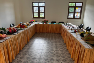 Aydınlık bir odada U şeklinde kurulmuş bir sergi masası. Masa, parlak, şeftali rengi bir kumaşla tamamen örtülmüş. Masanın üzerinde, hayvan, çiçek ve geometrik şekil gibi renkli origami figürleri yer alıyor. Masanın ön tarafında, sergilenen parçaların adlarını veya yaratıcılarını belirten küçük kartlar görünüyor. Arka planda, duvarda asılı iki dikdörtgen pencere doğal ışık sağlıyor. 