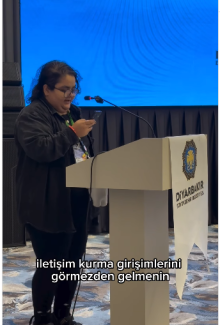 Çalıştayda kürsüde konuşma yapan, orta boylu, koyu renk saçlı ve gözlük takan Devrim Nesin görünüyor. Konuşmacı, siyah giysiler içinde ve hafifçe eğilmiş bir şekilde elindeki telefondan bir şey okuyor. Kürsü beyaz renkli ve üzerinde büyük, beyaz bir pankart sarkıyor; pankartta Diyarbakır Büyükşehir Belediyesi yazısı ile belediye logosu bulunuyor. Konuşmacının önünde, ekranın alt kısmında beyaz harflerle "iletişim kurma girişimlerini görmezden gelmenin" yazan bir altyazı görülüyor. Arka planda büyük, açık ma