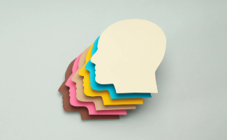 Bu görüntüde, farklı renklerdeki kağıtlardan kesilmiş, yan yana dizilmiş insan yüzü siluetleri bulunuyor. Siluetler üst üste gelerek bir yelpaze gibi duruyor. En üstte bej rengi bir siluet varken, altında sırasıyla mavi, sarı, pembe ve kahverengi tonlarında siluetler yer alıyor. Tüm siluetler aynı yöne, sola doğru bakıyor.