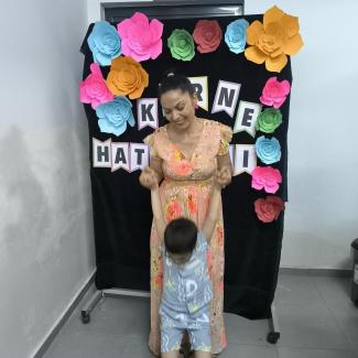 Resimde, siyah bir fonun önünde, gülen bir kadın var. Kadın, renkli çiçek desenli uzun bir elbise giymiş ve önünde, kollarından tuttuğu küçük bir çocuk diz çökmüş durumda. Arkadaki siyah fonda renkli kağıt çiçekler ve "KARNE HATIRASI" yazısı görülüyor.