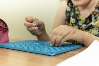 Çiçek desenli elbise giymiş bir Nazalin Selvi’nin elleri, mavi renkli bir plastik pegboard (delikli bir oyun veya eğitim tahtası) üzerinde küçük boncuk veya parçalar yerleştiriyor. Selvi’nin yüzü kısmen kadraj dışında kalmış. Masada ayrıca pembe bir çanta ve sağ alt köşede bir kâğıt var. Arka plan sade ve açık renkli.